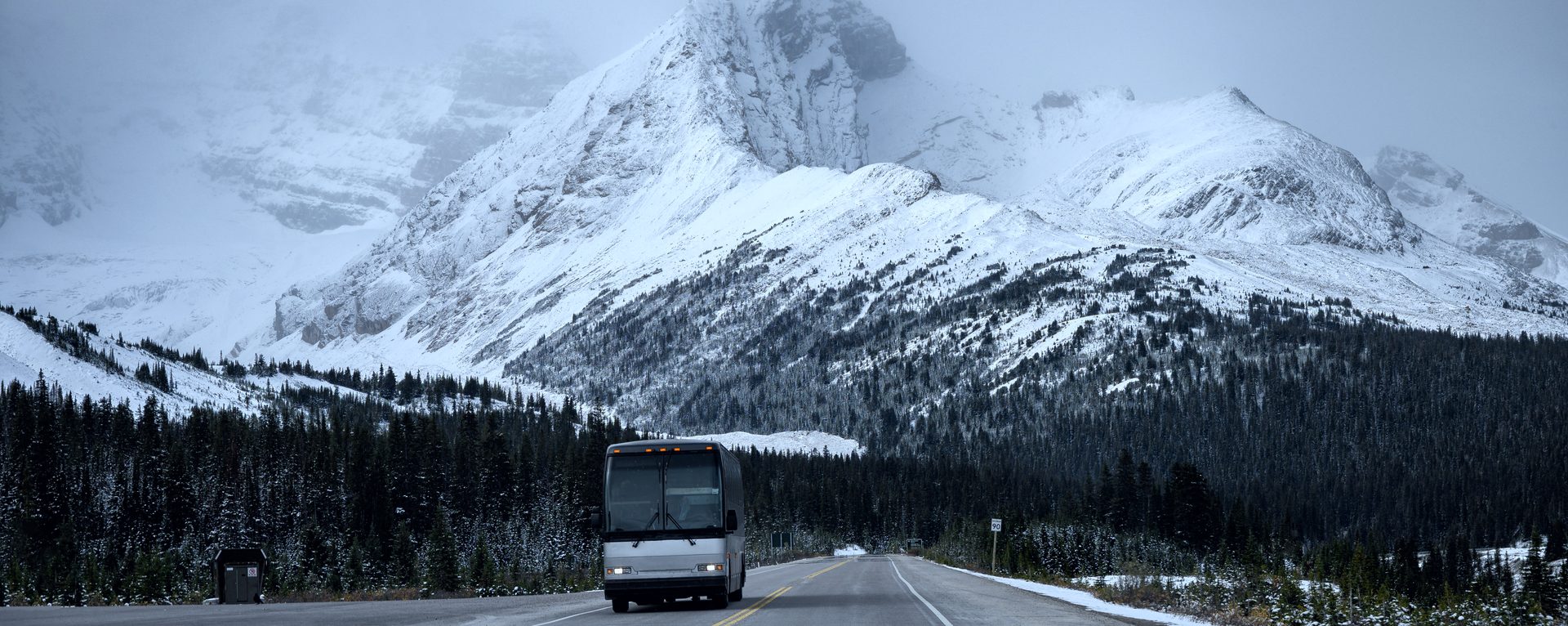 A motorcoach on a two-lane highway below a snow-covered peak