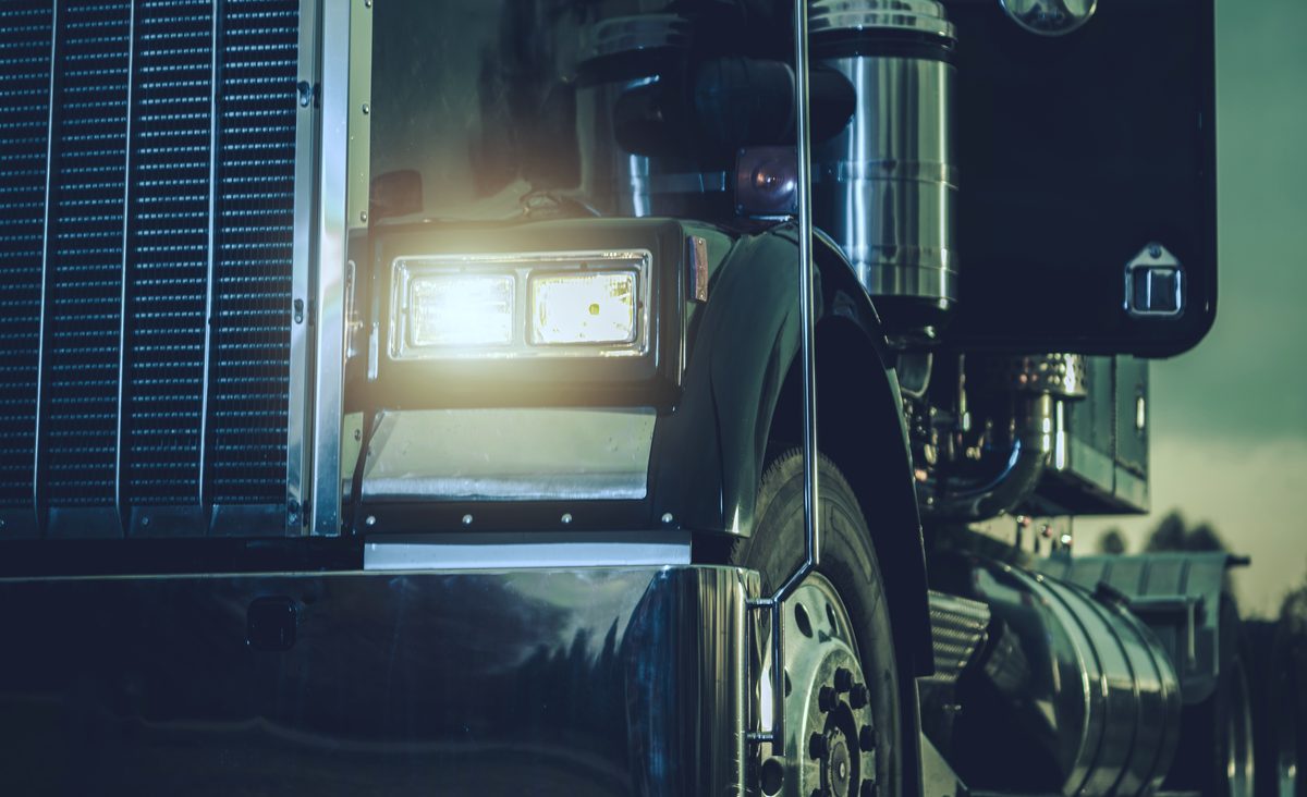 Headlamp, exhaust stack and fuel tank of a black semi truck at dusk
